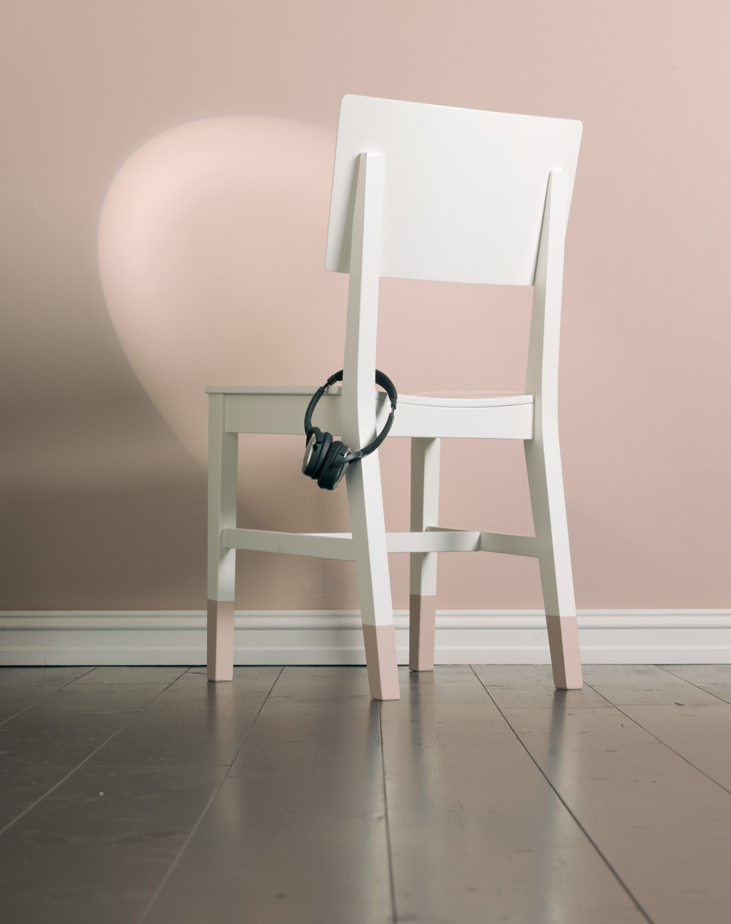 Hvit stol med rosa dyppede bein foran pastellrosa vegg, sorte hodetelefoner henger bak.