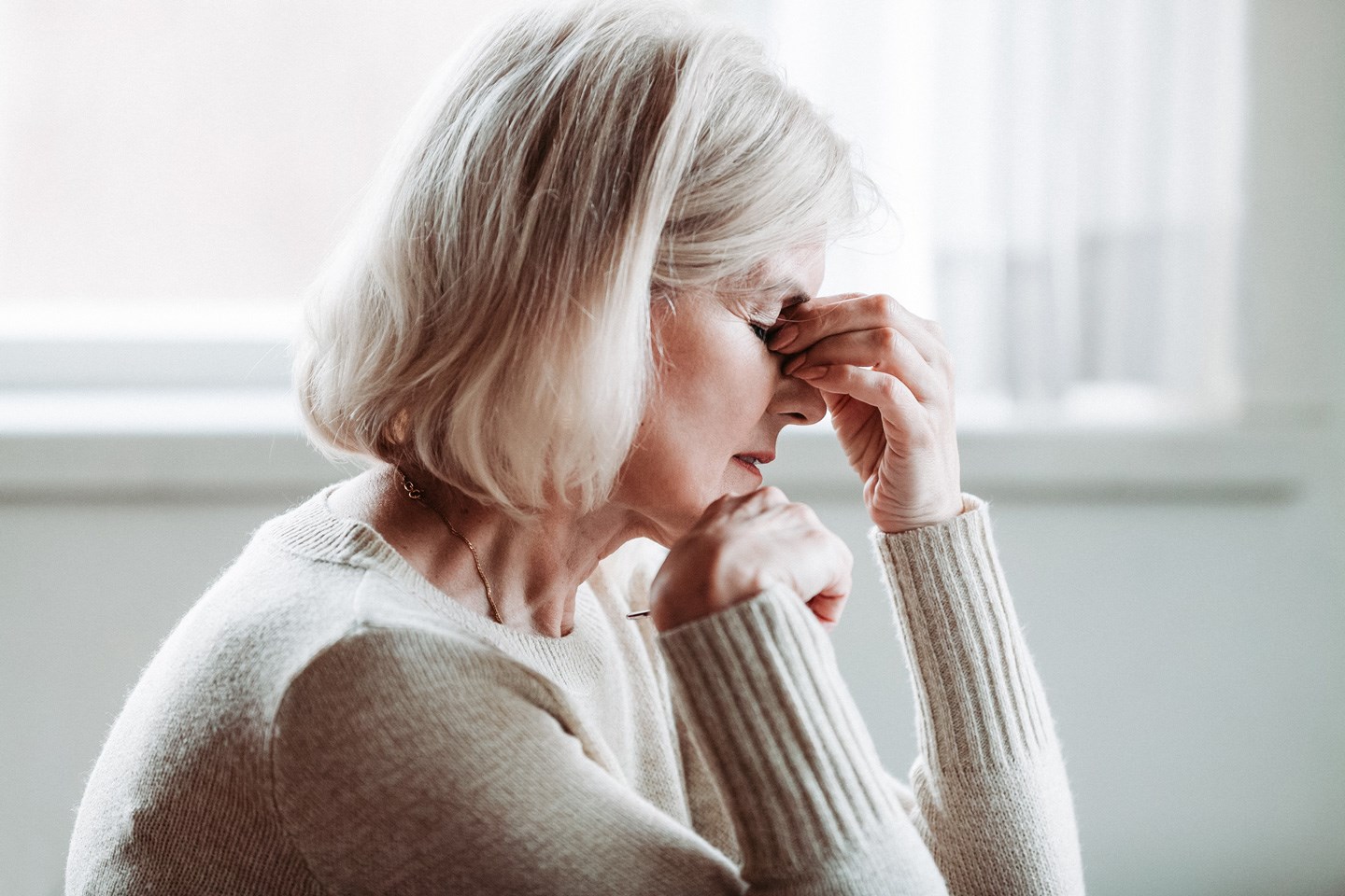Eldre person i beige strikkegenser sitter ved vindu i lyst rom med hånd mot kinnet.
