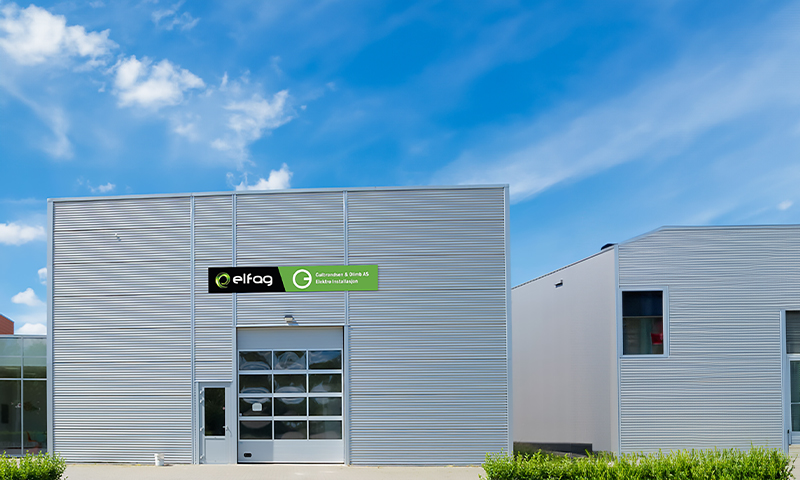 Grå industribygg med grønt Elfag-skilt, glassgarasjeport, blå himmel med skyer
