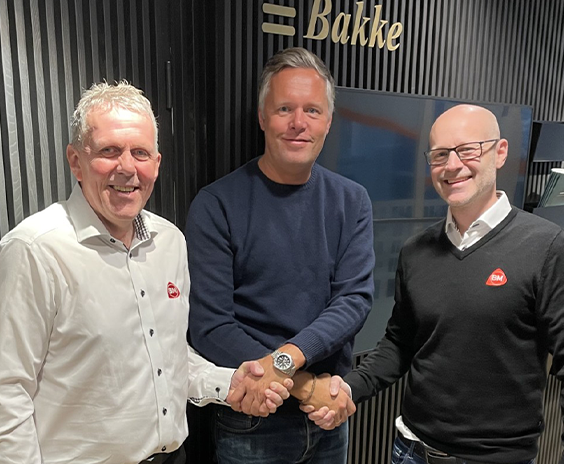 Tre menn i hvit, blå og svart genser håndhilser foran mørk panelvegg med Bakke-logo