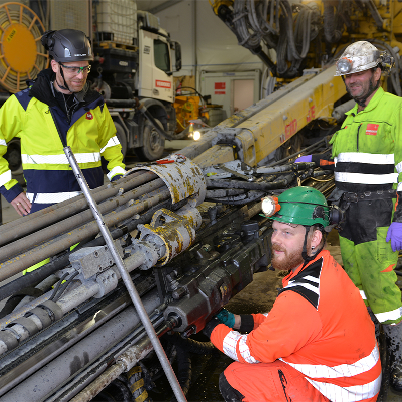 Arbeidere i verneutstyr ved en stor, gul boremaskin innendørs med stålrør og slanger.