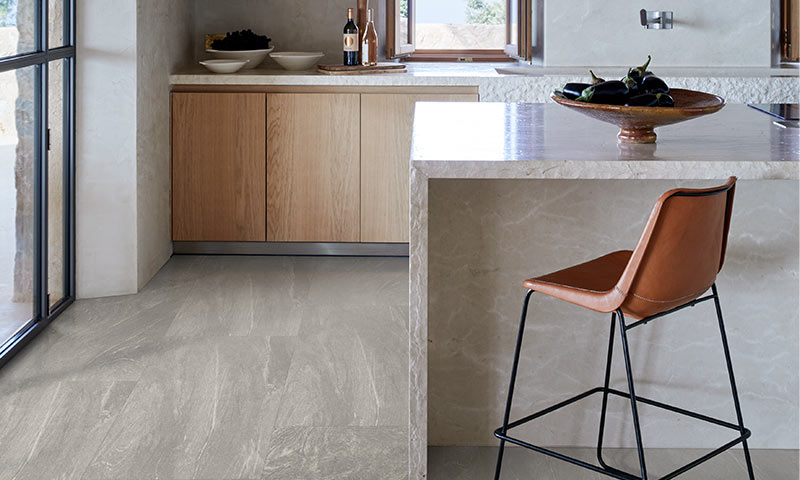 Moderne kjøkken med lys marmorøy, lyse trefronter, brunt skinnbarstol og auberginer i skål.