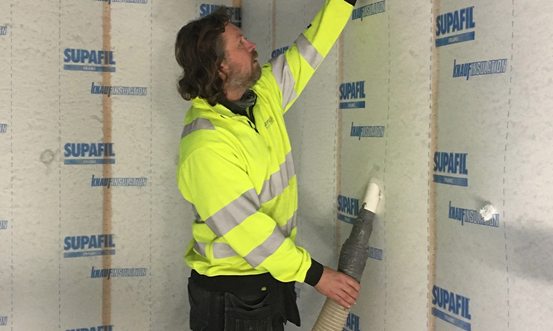 Arbeider i gul refleksjakke blåser SUPAFIL-isolasjon inn i trevegg.