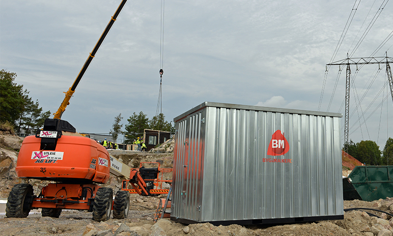 Byggeplass med oransje lift, sølvgrå container med rød BM-logo og kran under grå himmel