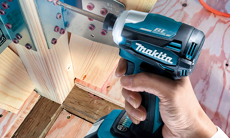 Hånd med blå-svart Makita skrutrekker bor i metallvinkel festet til lyst treverk