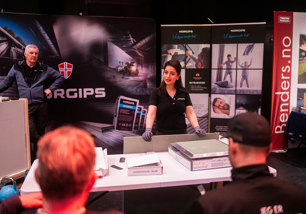 Kvinne i Norgips-uniform presenterer byggematerialer ved en stand på en messe, med publikum i forgrunnen.