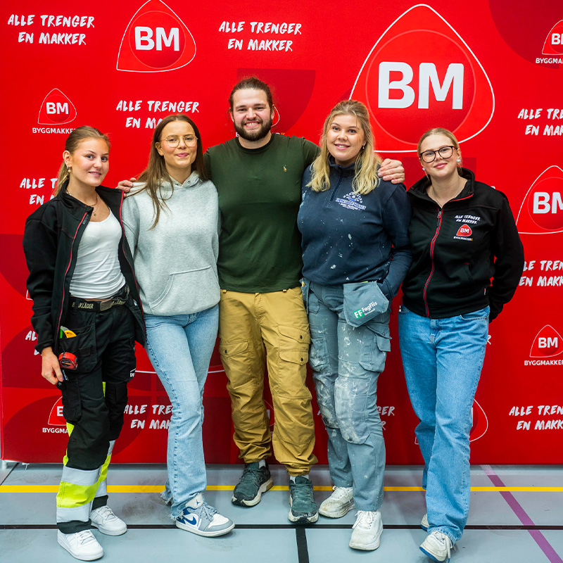 Fem personer i arbeidstøy og fritidsklær foran rød BM-vegg med «Alle trenger en makker»