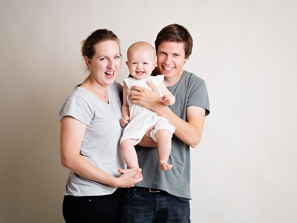 Foreldre i grå t-skjorter holder baby i hvit kjole foran lys nøytral bakgrunn.