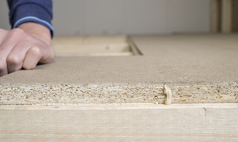 Hånd med blå erme presser sponplate på lys treramme med lim som pipler ut