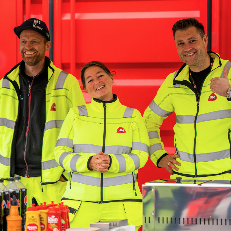 Tre personer i gul arbeidsjakke med refleks og BM-logo foran rød vegg, bord med brus og sauser.