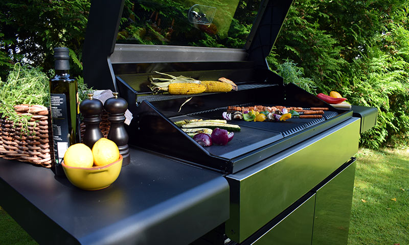 Åpen svart gassgrill i grønn hage med mais, grønnsaksspyd, pølse, rødløk, sitroner.