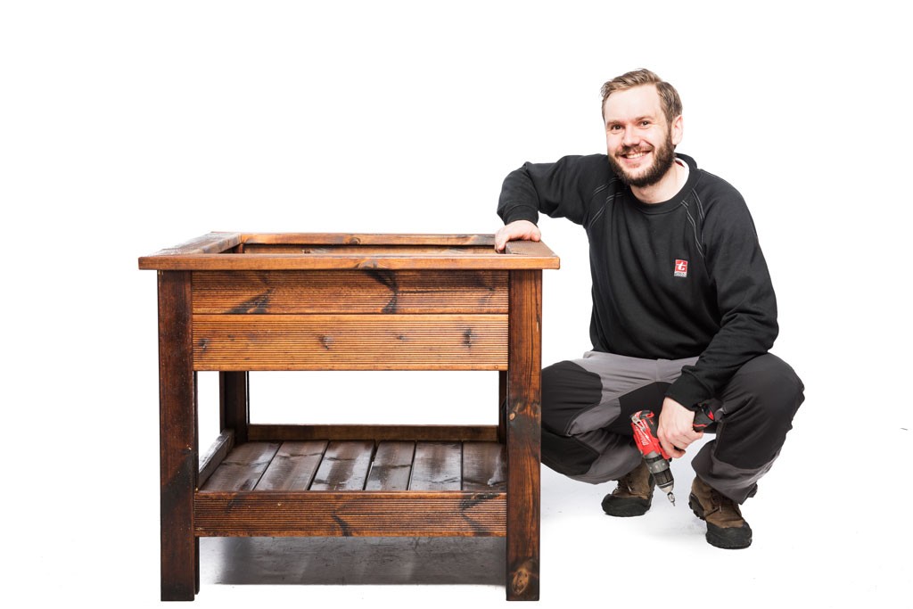 En mann i svart arbeidstøy knekker ned ved brunt trebord og holder en rød drill på hvit bakgrunn.