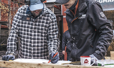 To håndverkere i rutete skjorte og svart jakke måler en treplanke ved byggeplass med sveisesprut.