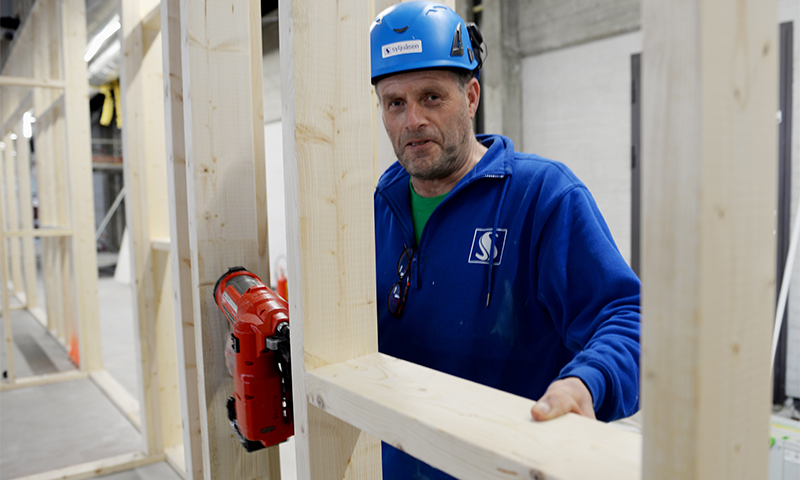 Arbeider i blå hjelm og genser fester lys treramme med rød spikerpistol.