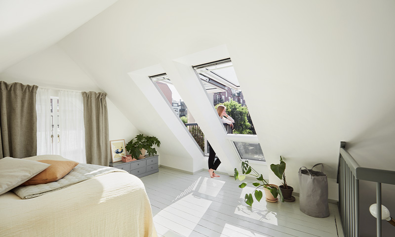 Lyst loftsoverom med skråtak, hvite vegger, store takvinduer, grønne planter og kvinne ved vindu.