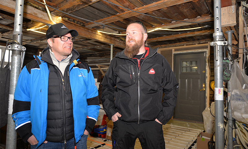 To menn i blå og svart jakke står i kjeller under oppussing med synlige trebjelker.