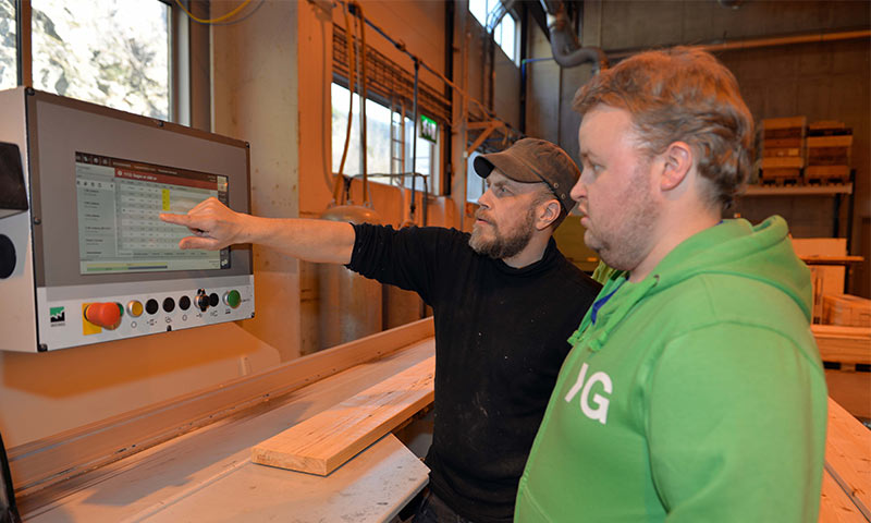 To menn ved kontrollpanel i treverksted, den ene peker på skjermen ved sagmaskin.