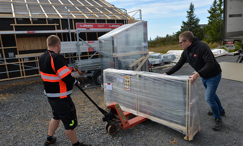 To arbeidere frakter folieinnpakket pall på rød tralle ved rammebygget trehus.