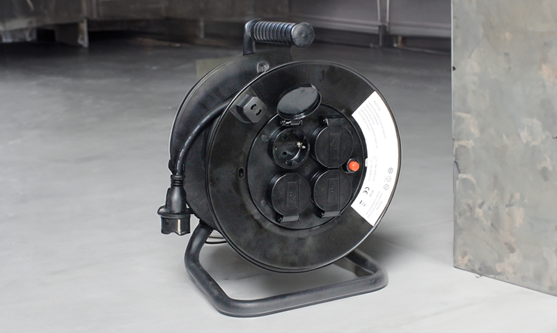 Svart kabeltrommel med fire stikkontakter og rødt knapp på grå industrigulv.