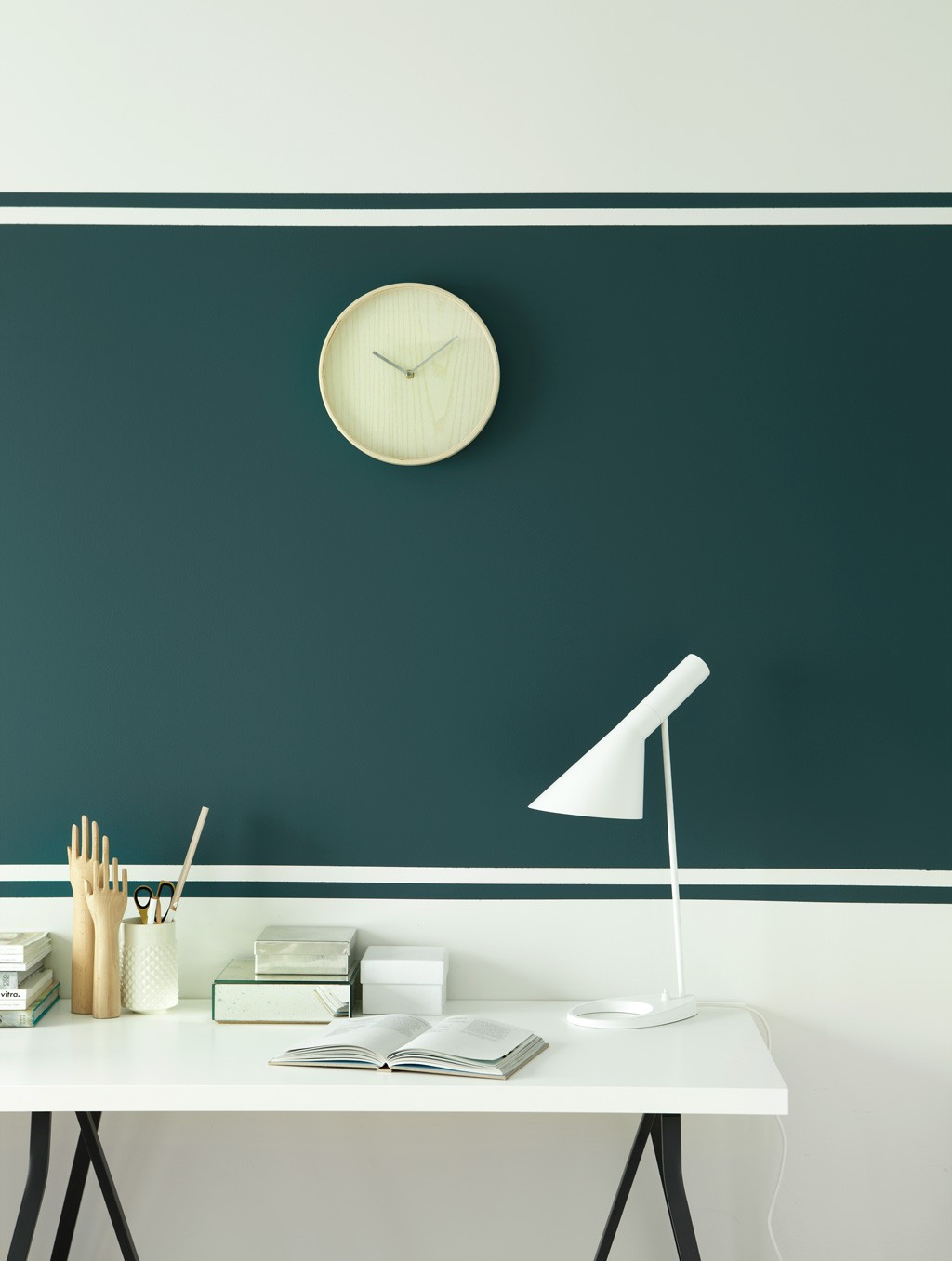 Hvitt skrivebord med lampe, åpen bok penneholder mot mørk grønn vegg med hvite striper og treklokke