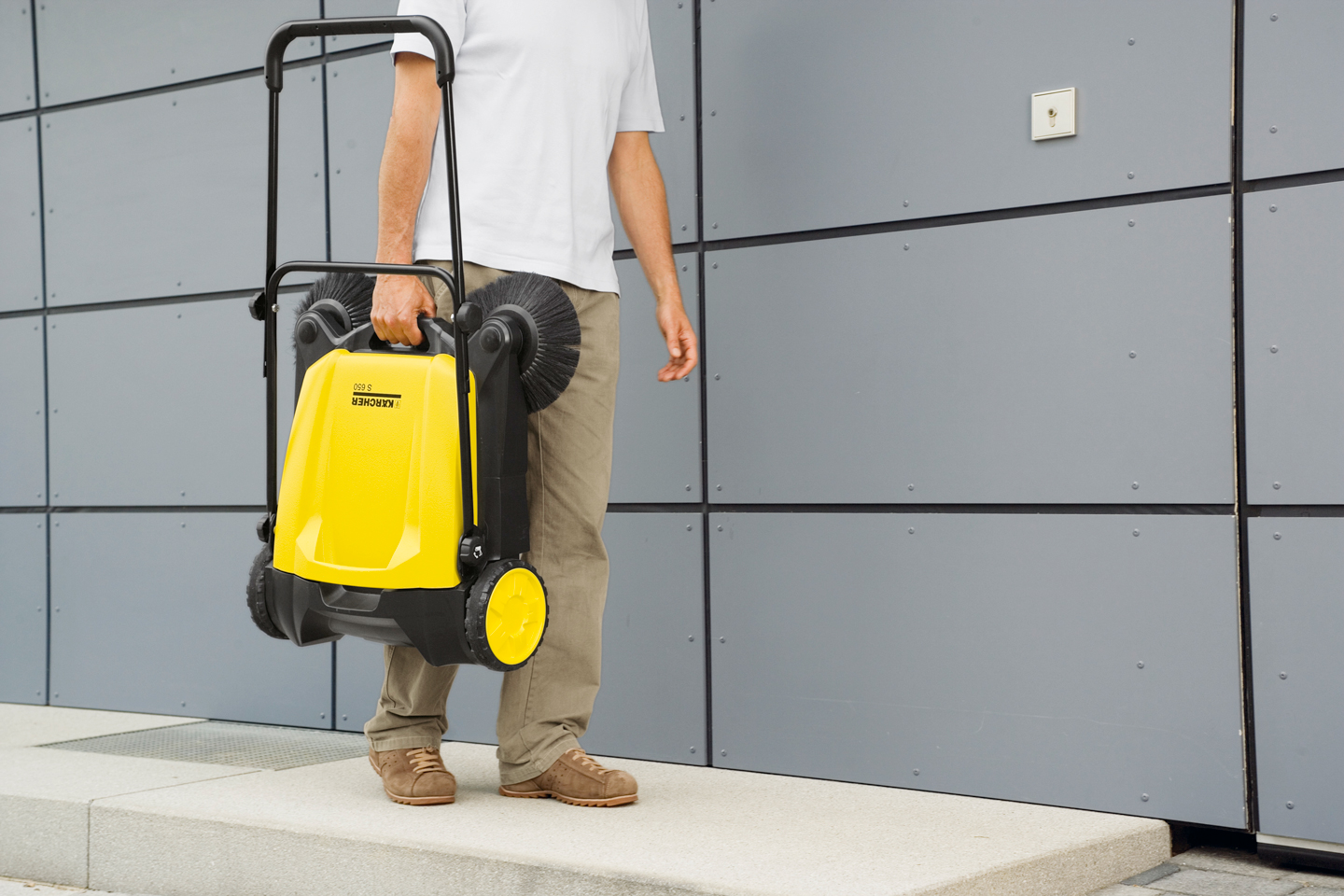 Person bærer gul-svart feiemaskin med hjul foran grå vegg.