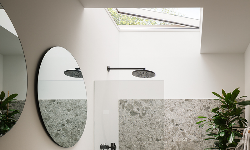 Moderne lyst baderom med takvindu, svart regndusj, grå terrazzovegg, rundt speil og grønn plante.