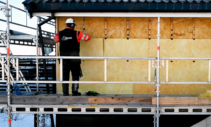 Arbeider med hvit hjelm og rød-sort vest monterer gul isolasjon på husvegg fra stillas