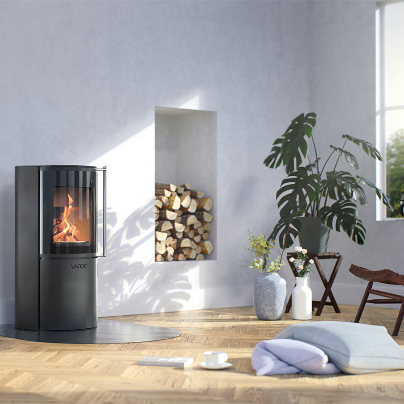 Lyst rom med svart vedovn i bruk, innebygd vednisje, grønne planter og puter på tregulv.