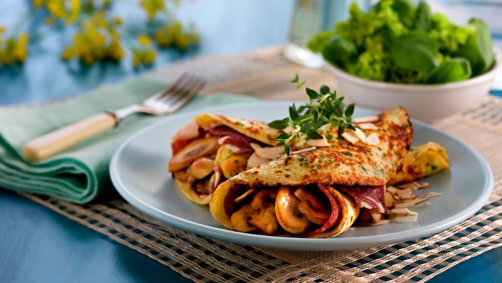 Blå tallerken med gylne urtepannekaker fylt med sopp og skinke, toppet med mandelflak og timian