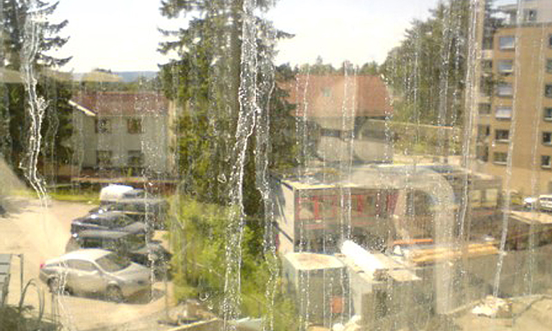 Utsikt gjennom regnstriper på vindu mot parkeringsplass med biler, grantrær og leilighetsbygninger.