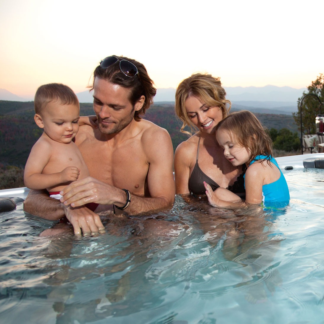 Foreldre med to barn i boblebad foran fjellkjede i solnedgang.