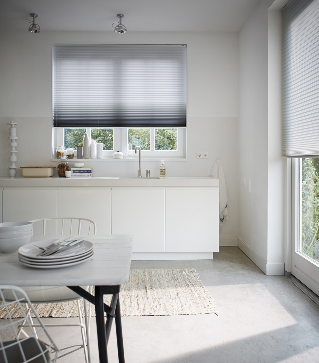 Lyst hvitt kjøkken med vindu dekket av grå-hvit plisségardin, benk og bord med tallerkener