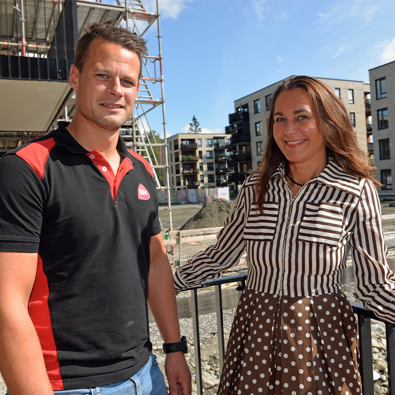 En mann i svart-rødt arbeidstøy og kvinne i brun-hvit stripet kjole foran byggeplass med stillas.