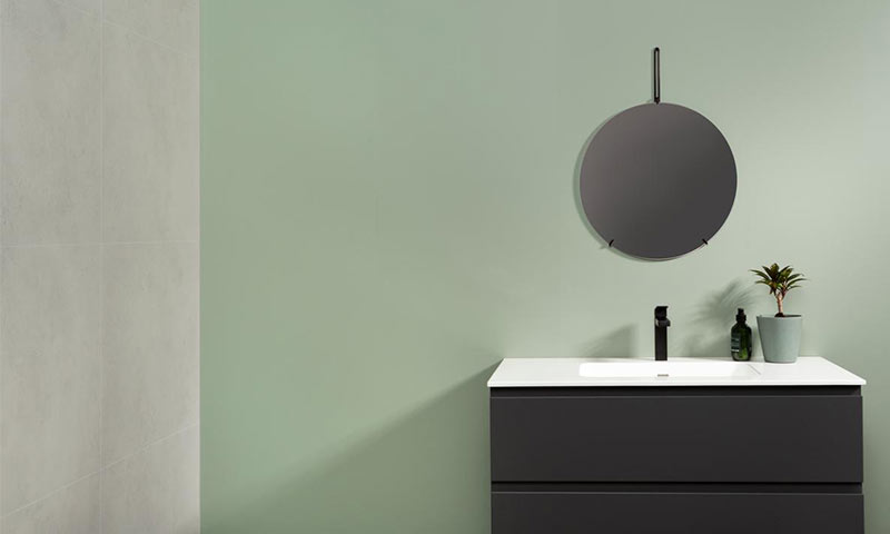 Minimalistisk baderom: svart servant, hvit benkeplate, rundt speil, grønn vegg og plante