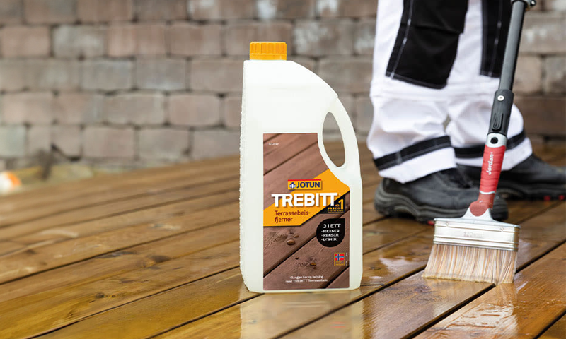 Hvit Jotun Trebitt-kanne med oransje kork på vått brunt terrassegulv mens hånd med pensel renser