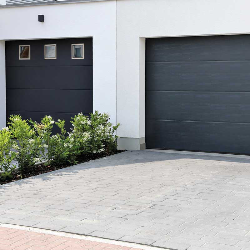 Moderne hvitt hus med to mørke garasjeporter, lys grå oppkjørsel og grønn hekk.