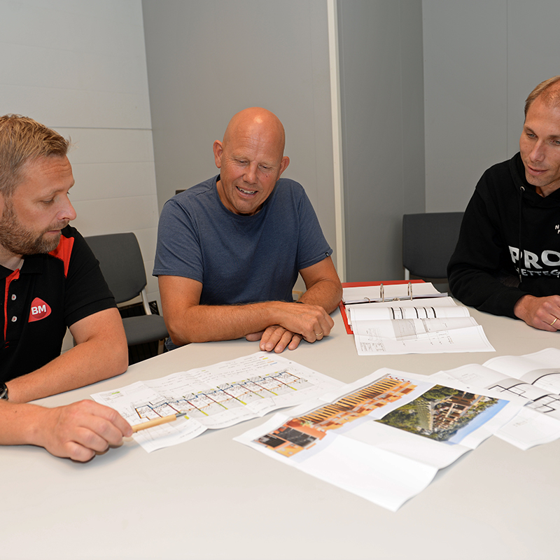 Tre personer ved et bord med arkitekttegninger, byggeplaner og foto av hus.