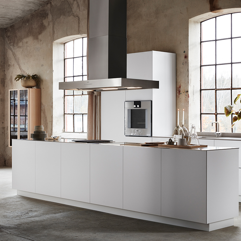 Hvit kjøkkenøy med svart ventilator, innebygd ovn, betongvegger og store vinduer.