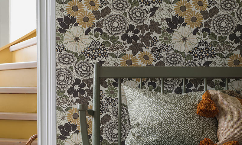 Grønn benk med mønstrete puter foran brunbeige blomstrete tapet og gul trapp i bakgrunnen.