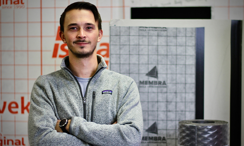 Person med armene i kors i grå fleecejakke foran hvit rutenettvegg med oransje logo og byggevarer.