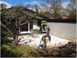 To håndverkere støper hvit betongplate i hage foran det grå huset under blå himmel