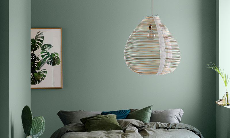 Lyst soverom med mintgrønne vegger, flettet lampe, uredd seng med grønne puter og plantebilde