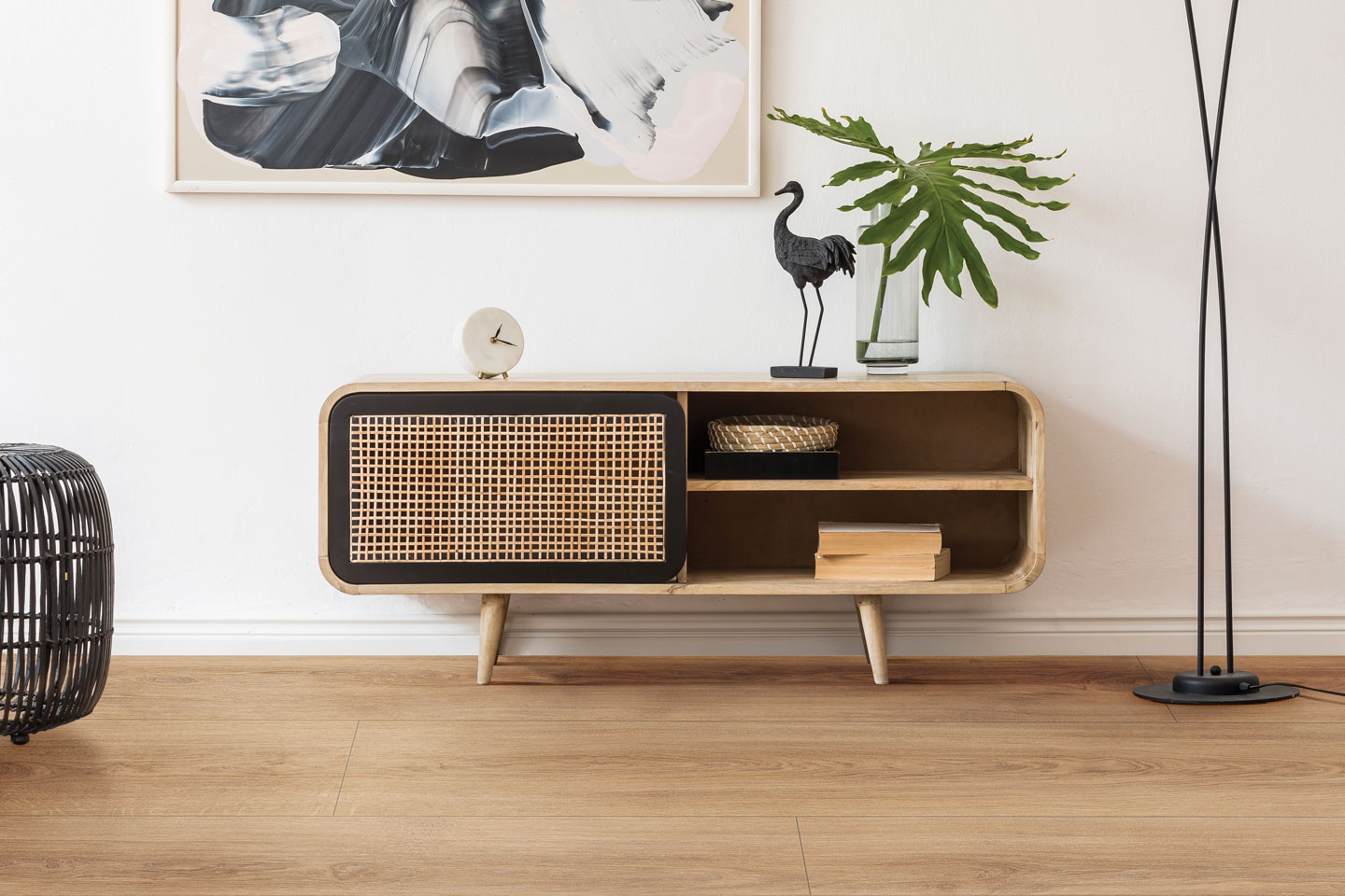 Stilrent rom med eikegulv: lys tre-sjenk med rattanfront, grønt blad, svart lampe, sort-hvitt bilde.