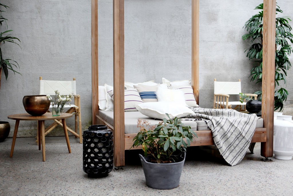 Ute-lounge med trebaldakin, hvite puter, stripete pledd, bambusstoler og grønne planter.
