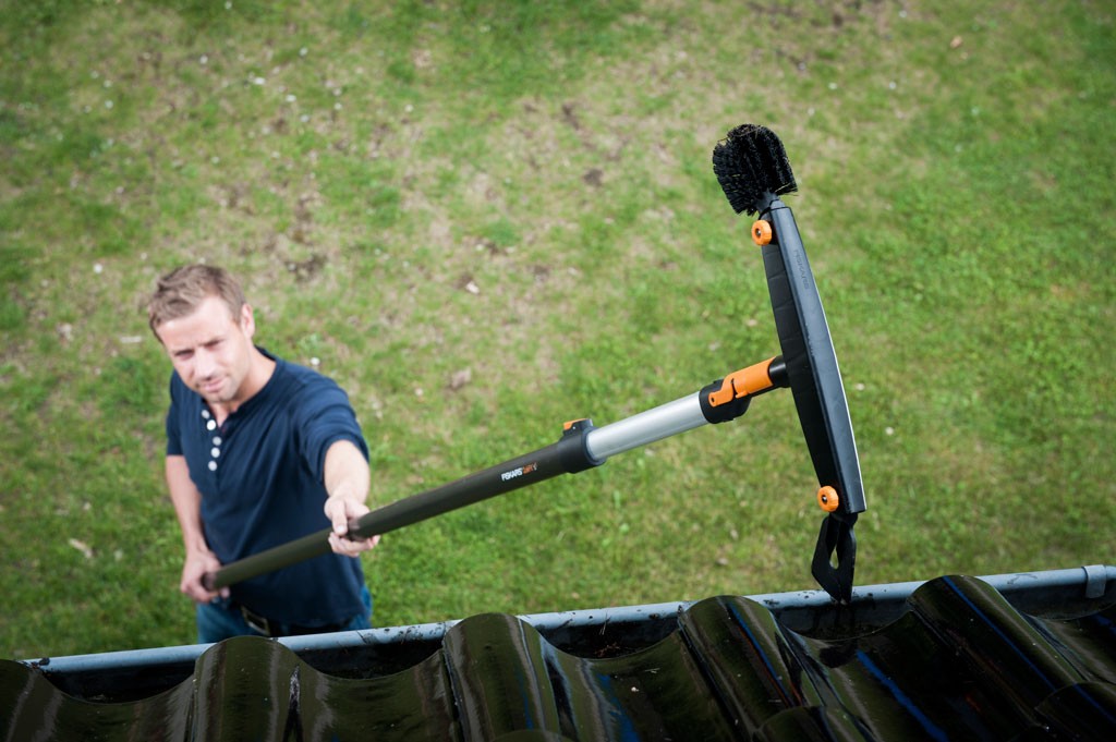 Mann står på bakken og rengjør svart bølgepapp-takrenne med teleskopkost med oransje detaljer.