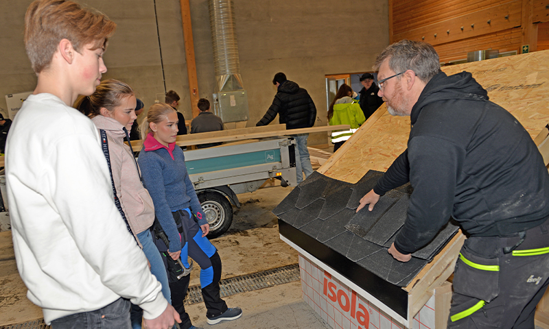 Håndverker viser svarte takshingel til ungdom i lyst verksted med trebjelker.