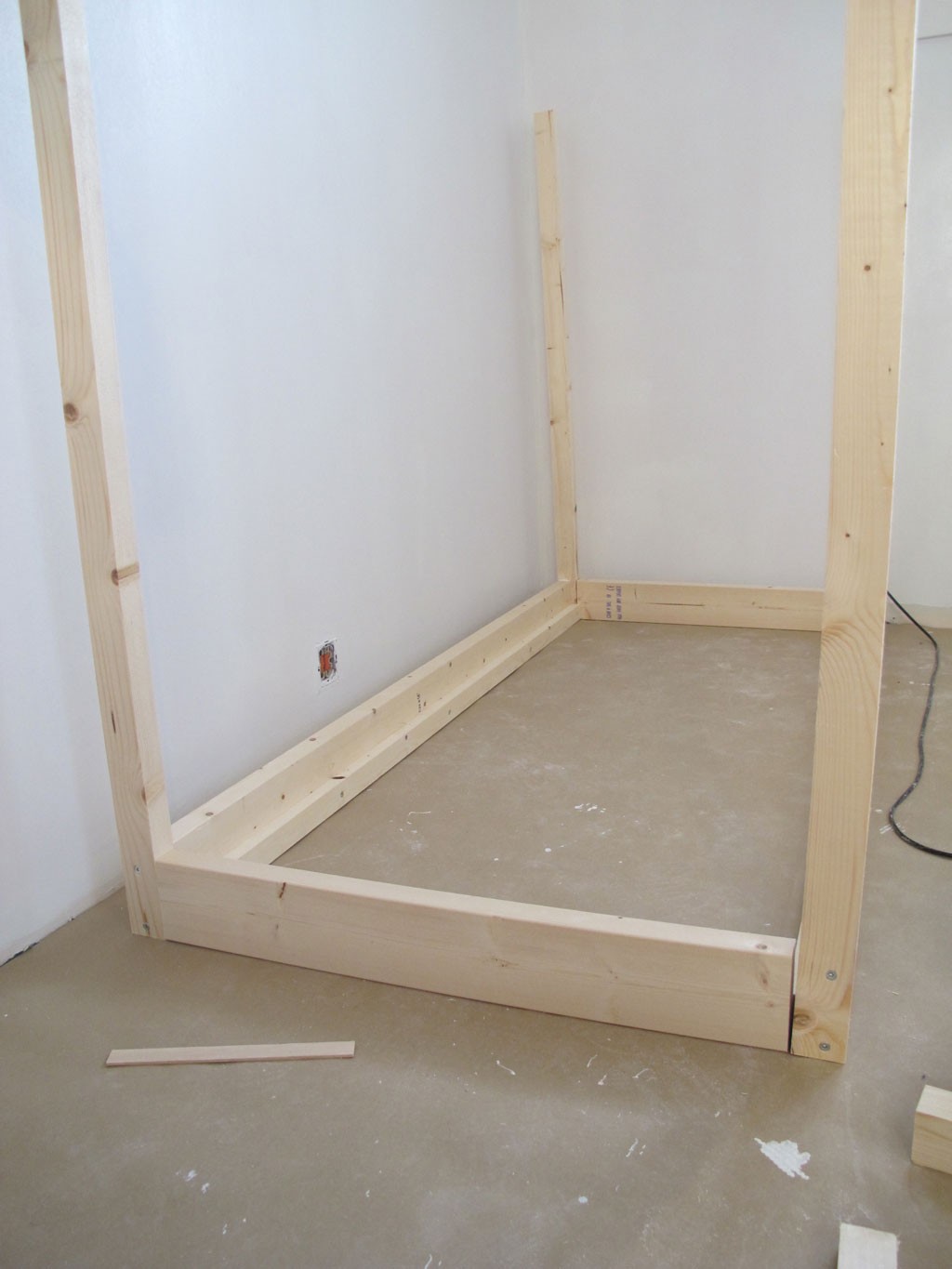 Uferdig lyst treramme montert mot hvite vegger på grått betonggulv.