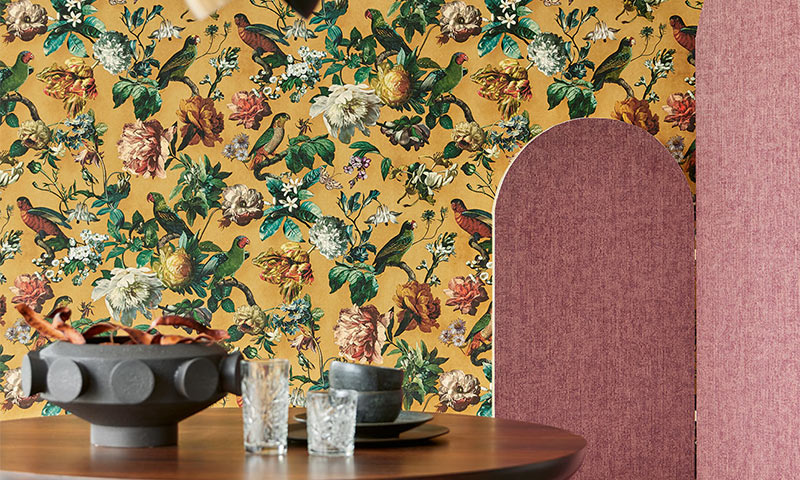 Keramikk og glass på trebord mot sennepsgul blomster- og fugletapet, ved rosa stoffskjerm.
