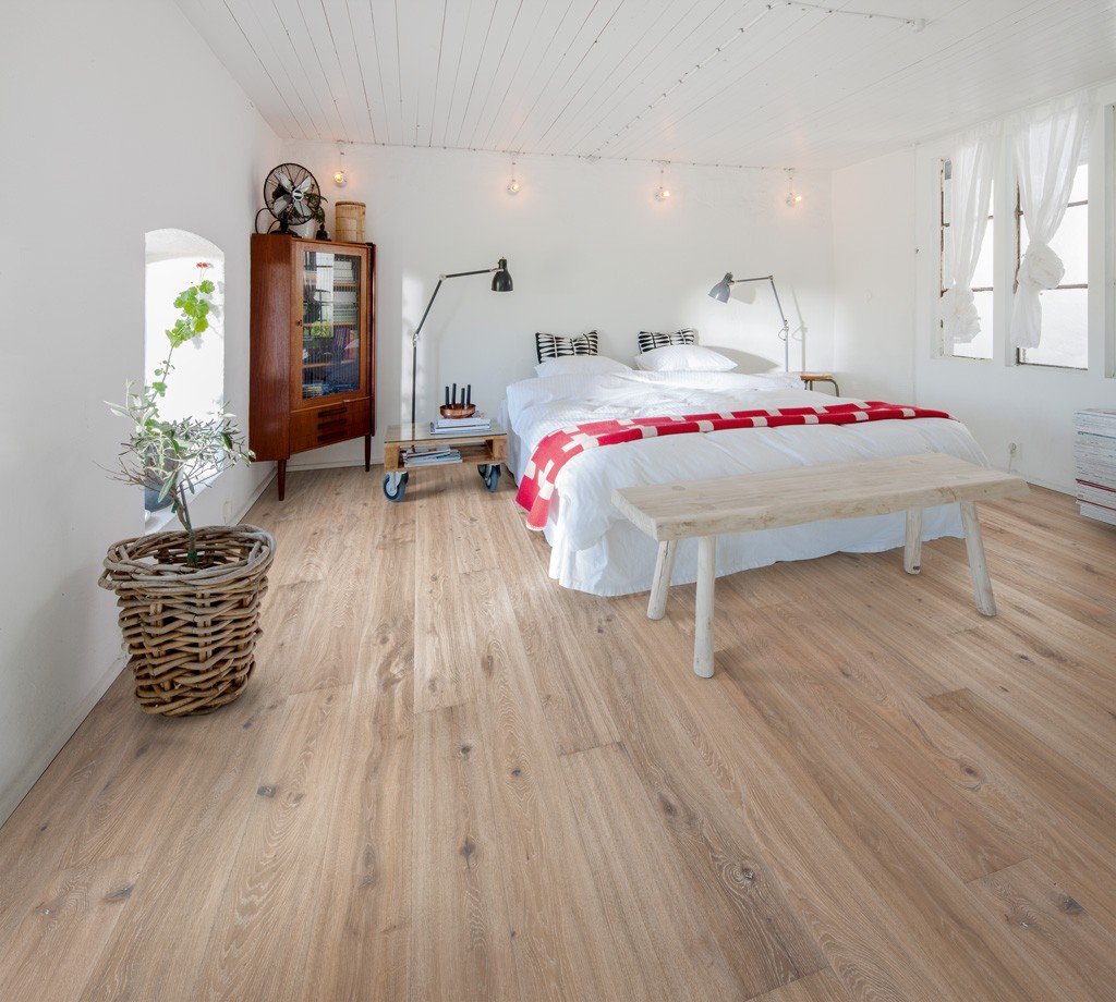 Lyst loftsoverom med hvite vegger, eikegulv, seng med rødt pledd, gamle møbler og grønne planter.