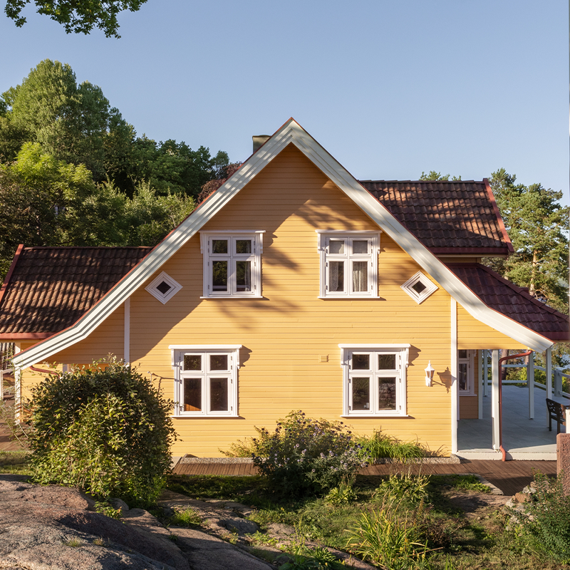 Gult trehus med rødt tegltак, hvite vinduer og veranda, omgitt av grønn vegetasjon og blå himmel.